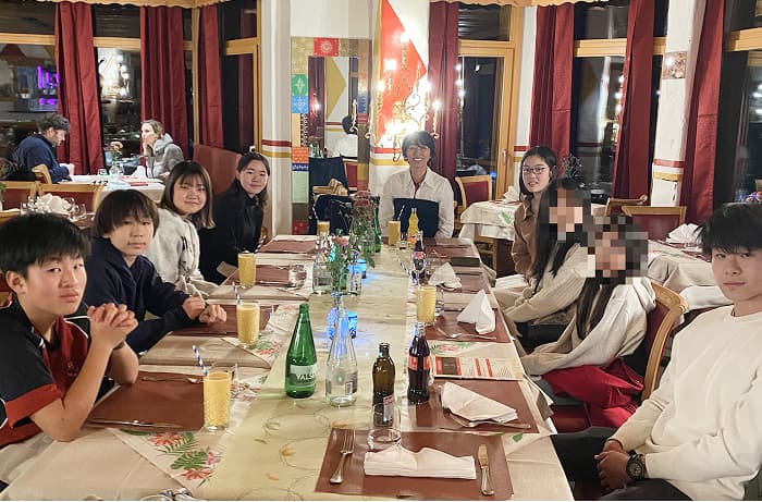 夜のレストランの中で長いテーブルに日本人留学生が8人、日本人の現地スタッフが1人座っています。テーブルには飲み物や食器が並べられ、食事会が始まろうとしています。人物は皆撮影者の方を向いており、にこやかに笑顔を向けています。